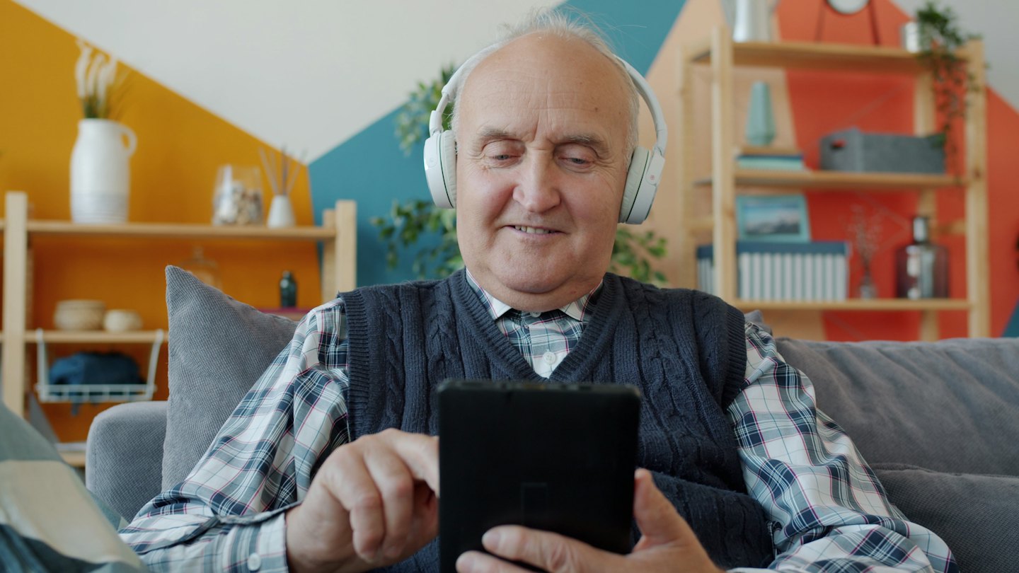 ヘッドフォンをつけた年配の男性がソファでタブレットを使いながら微笑んでいる。